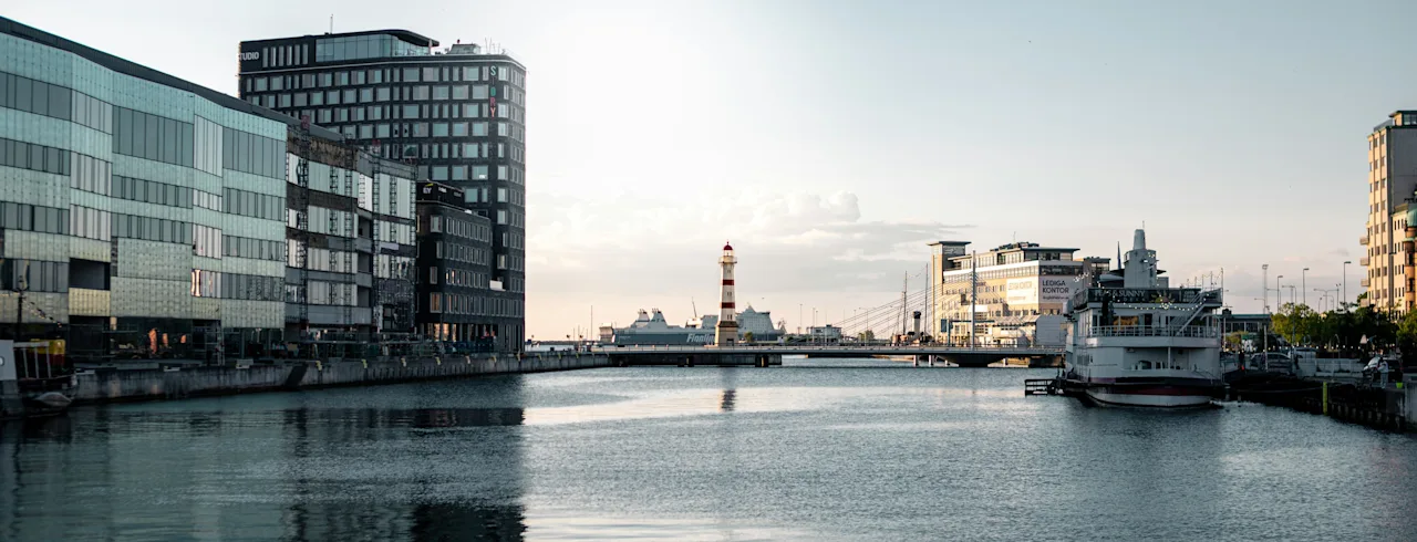 Malmö havneområde med moderne bygninger, et rødt og hvitt fyrtårn, og en båt. Synlig tekst inkluderer STUDIO og LANGGA KONTOR.