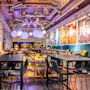 Modern dining space with chairs and tables, vibrant artwork on the walls, and large, spherical lights overhead. Industrial decor with exposed ducts and colorful ambiance.