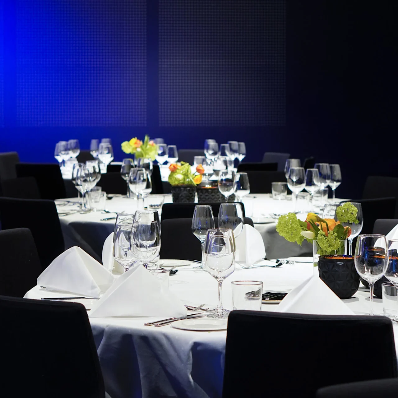 Tables set elegantly with white tablecloths, folded napkins, wine glasses, and floral centerpieces, in a dimly lit dining environment with a blue-lit background.