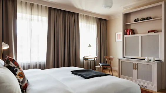 Seng med mønstrede puder og sort tæppe på hvidt sengetæppe, i et hyggeligt rum med brune gardiner, et lille bord, en lampe, og et beigefarvet skab fyldt med bøger og dekoration.