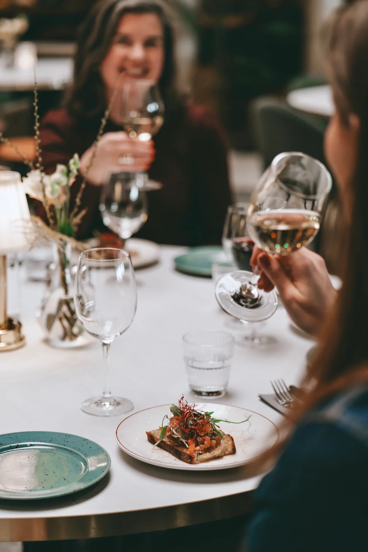 A plate with toast topped with greens sits on a table. A person holds a wine glass, with other diners blurred in the background, suggesting a social meal setting.