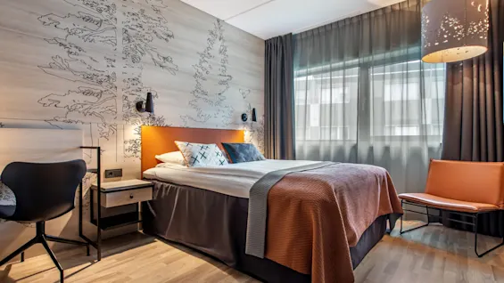 A neatly made bed with orange and gray bedding stands in a modern room with wooden floors, artistic wall drawings, and soft lighting from a large lamp.