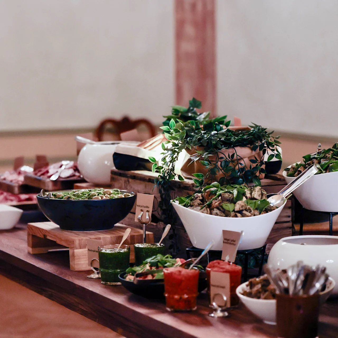 A buffet table features assorted dishes in white bowls, including salads and sauces, accompanied by serving utensils. It is set in an elegant dining room with pink-accented walls.