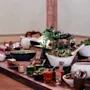 A buffet table features assorted dishes in white bowls, including salads and sauces, accompanied by serving utensils. It is set in an elegant dining room with pink-accented walls.