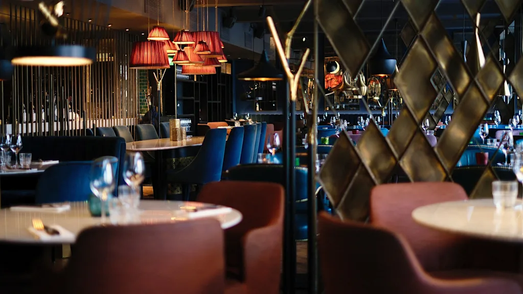 Elegante restaurantborde dækket med glas og tallerkener, omgivet af farverige stole. Varm belysning fra hængende lamper skaber en hyggelig atmosfære, med geometrisk dekoration i baggrunden.