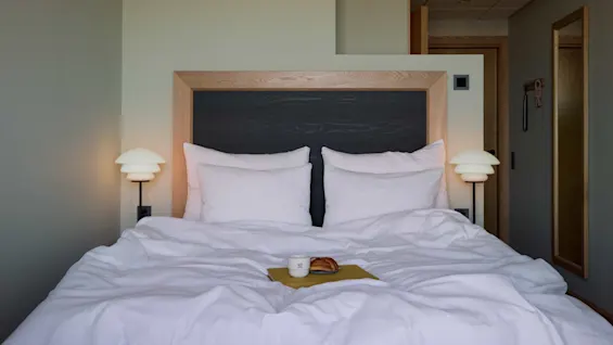 A neatly arranged bed with white pillows and lamps sits in a minimalistic room. A coffee cup and croissant are on a napkin. Text on the cup reads "MB."