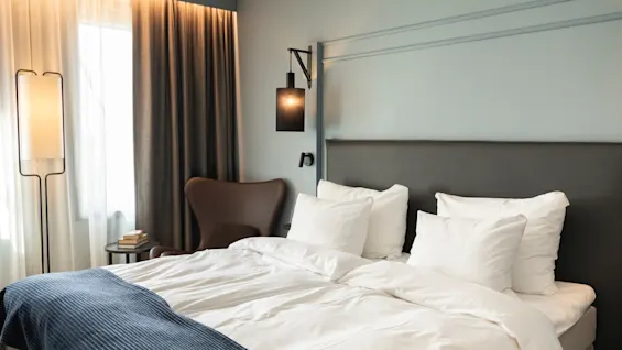 Et koselig hotellrom med en seng med hvitt sengetøy og et blått pledd, en lenestol og varm belysning.