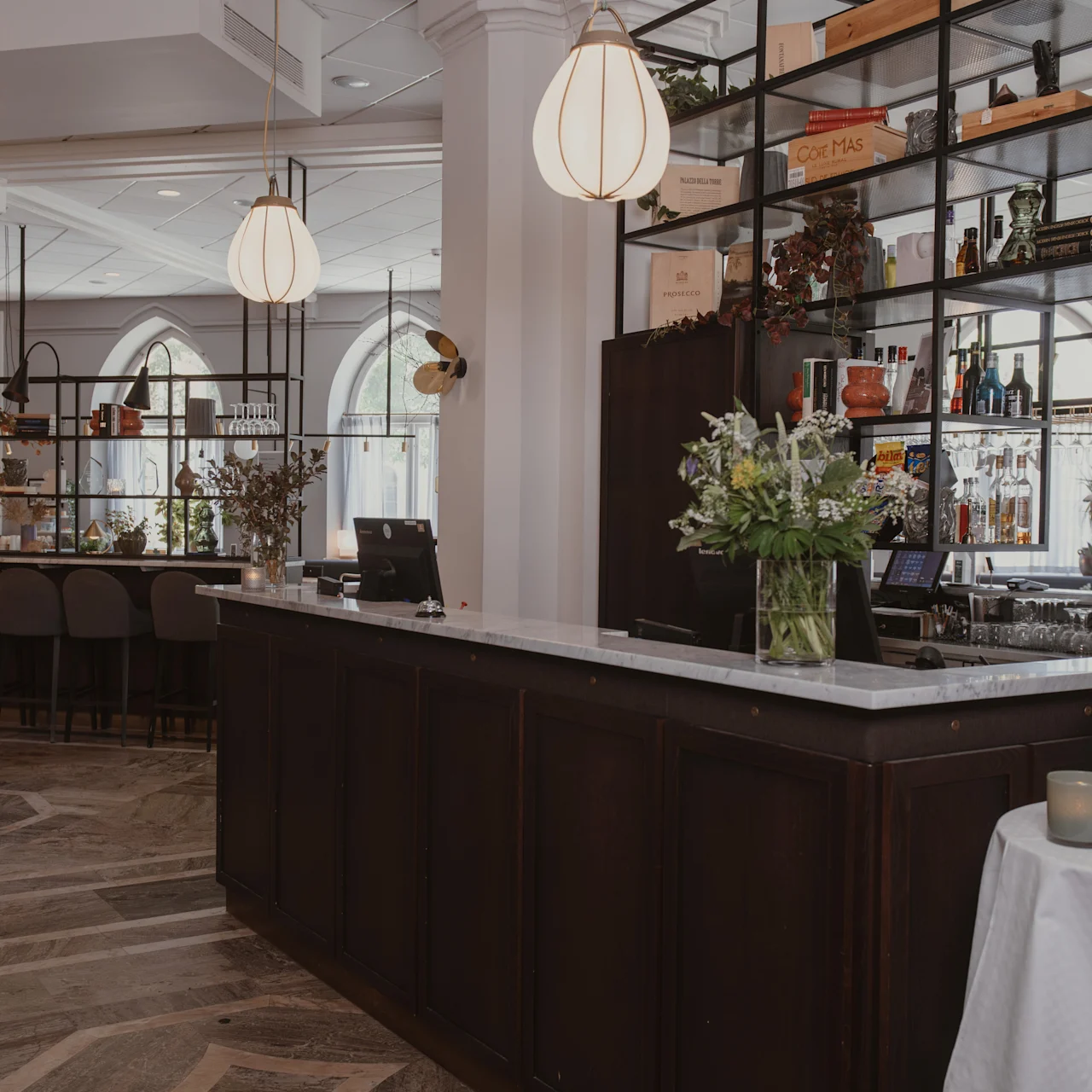 En bardisk prydd med blomvaser, i en elegant restaurangmiljö med moderna taklampor, höga hyllor med böcker och flaskor, och stiliga sittplatser.