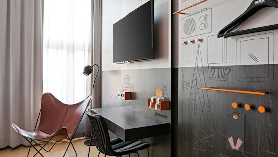 En veggmontert TV i et moderne rom, over et skrivebord med to stoler. En gulvlampe og skinnstol er i nærheten, med minimalistisk dekor på veggene.