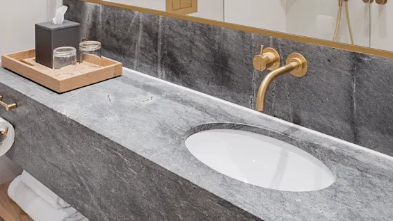 A modern bathroom with a marble countertop features a gold faucet above a white sink, with a mirror reflecting a shower area. A wooden tray holds glasses and tissues.