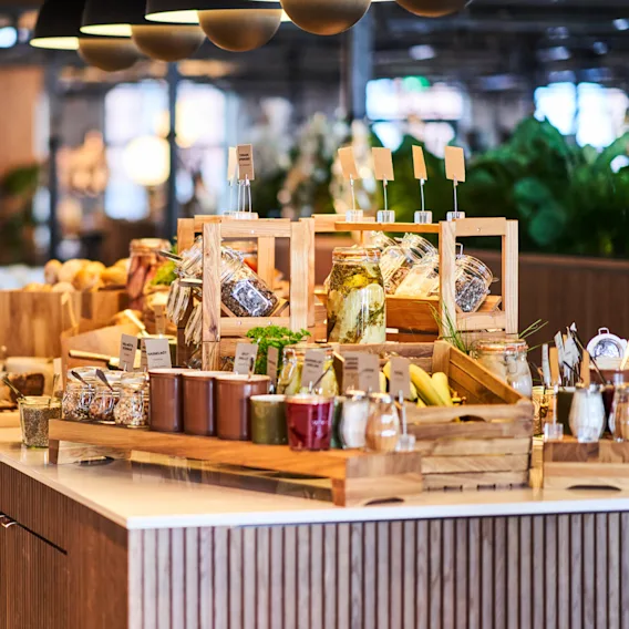 Ett buffébord innehåller burkar med pickles, kryddor och örter på en trädisk i en väl upplyst restaurang. Text på etiketter lyder: ”Nudelsalat,” “Parmesan,” och “Olivenöl.”