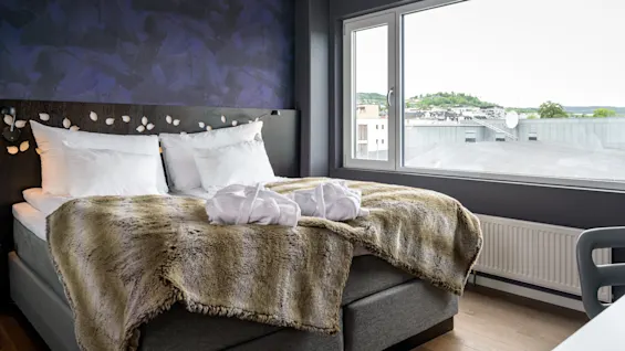 A neatly made bed with white pillows and a fur blanket sits beside a large window overlooking rooftops and greenery, in a room with dark blue walls.