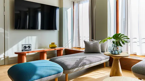 Et vægmonteret TV i en lys stue, kompletteret med en moderne grå sofa, blå puf, glasbord med en plante og lysfiltrerende gardiner.
