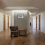 Hotellikäytävä, jossa on puulattiat, useita ovia, viihtyisä nojatuoli, pieni pöytä ja kehystetty kuva seinällä.