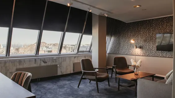 Et moderne hotell loungeområde med store skråstilte vinduer som tilbyr byutsikt, møblert med komfortable sitteplasser og et salongbord.