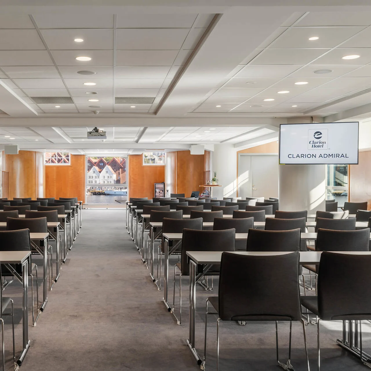 Rækker af stole vender mod et lærred i en konferencelokale. To skærme viser teksten "Clarion Hotel, CLARION ADMIRAL." Store vinduer bagved giver naturligt lys.