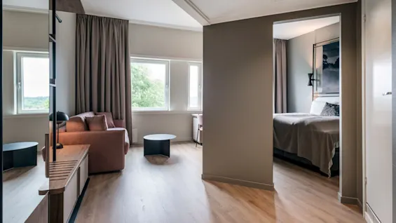 Et koselig hotellrom har en brun sofa og et lite bord ved store vinduer med utsikt over trær. En seng er delvis synlig gjennom en tilstøtende døråpning.