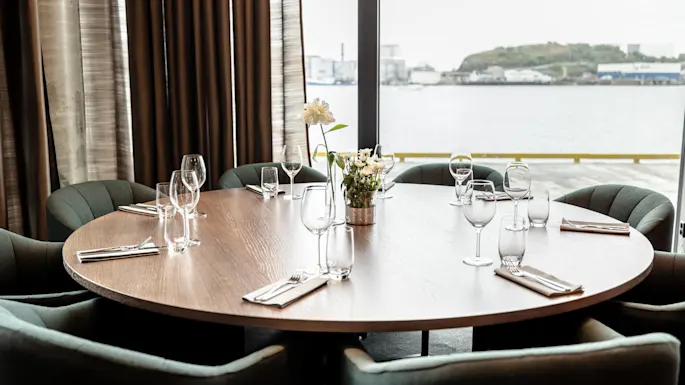 Round table set with wine glasses and napkins, surrounded by upholstered chairs, in a dining room with large windows overlooking a waterfront and industrial buildings in the distance.