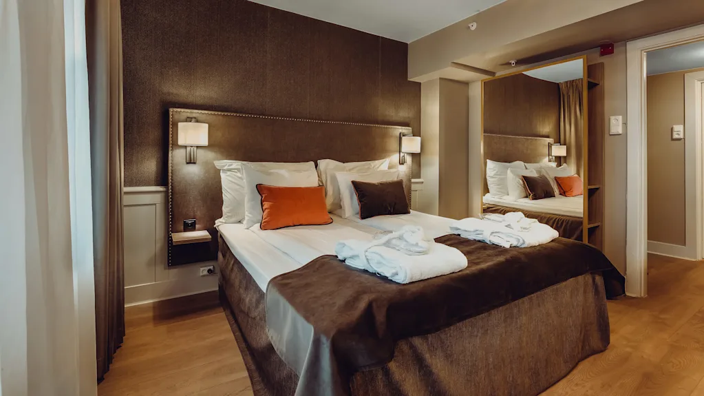 En pænt redt seng med hvidt sengetøj, akcentpuder og håndklæder i et godt oplyst hotelværelse.