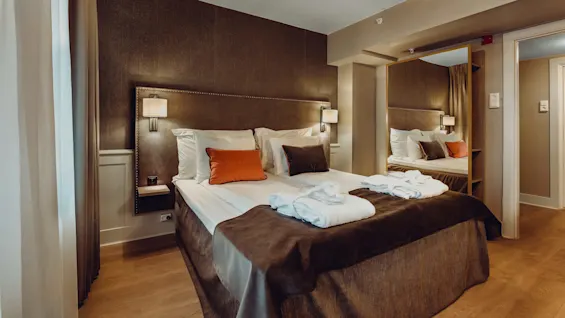 A neatly made bed with white linens, accented pillows, and towels in a well-lit hotel room.