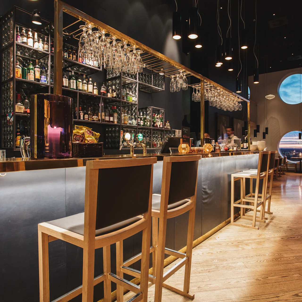 En bardisk kantet med høje træstole, glas hængende ovenover, og en række spiritusflasker bagved. Cirkulære vinduer med udsigt over en solnedgang, der oplyser restaurantens moderne indretning.