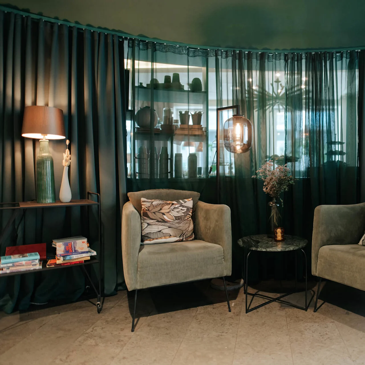 Two green armchairs with patterned pillows, a small marble table, and a shelf with decorations in a room with sheer curtains.