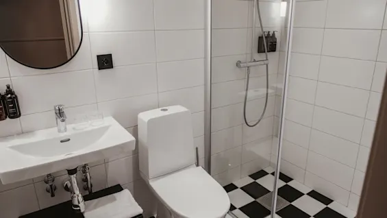 Ett rent, modernt badrum med vita kakelplattor, handfat, toalett och en dusch med svartvitt rutigt golv.