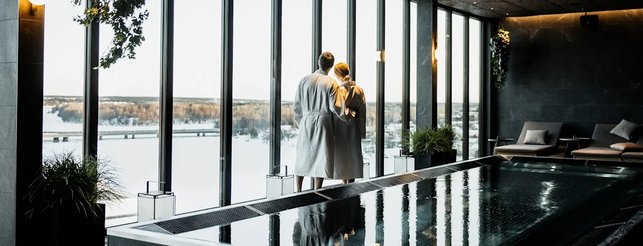 Two people in white robes stand by large windows, gazing outside. Indoors, a tranquil spa environment surrounds them with plants and lounge chairs. A rectangular pool reflects the scene.