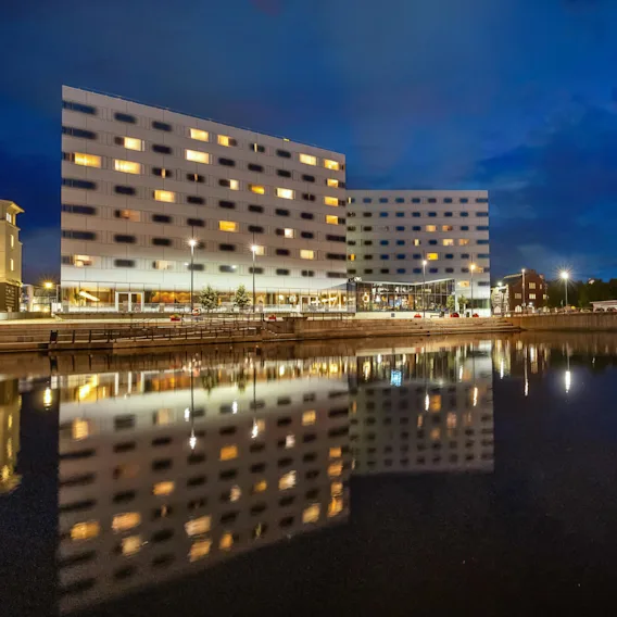 Store bygninger med opplyste vinduer gjenspeiles i den rolige vannkanten om natten; bygningen til høyre viser "ROCKHEIM" med lyse røde lys på fasaden.