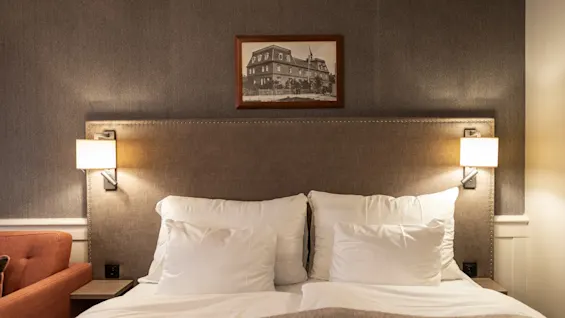 A cozy hotel room at Home Hotel Grand Bodø with a neatly made bed, a padded headboard, and warm lighting from wall lamps. A framed picture hangs above the bed.