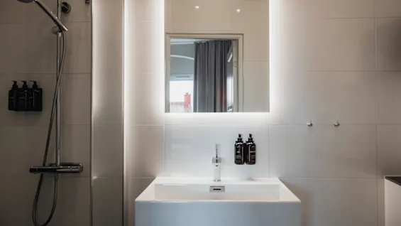 Et moderne badeværelse med bruser og håndvask. Spejlet over vasken er baggrundsbelyst. Dispensere til SHAMPOO, BALSAM, BODY WASH, HÅNDSÆBE, LOTION er synlige.