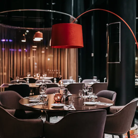 Ett runt middagsbord dukat för en måltid, omgiven av stoppade stolar. Ovanför ger en stor röd lampa ljus. Miljön är en svagt upplyst, modern restaurang.