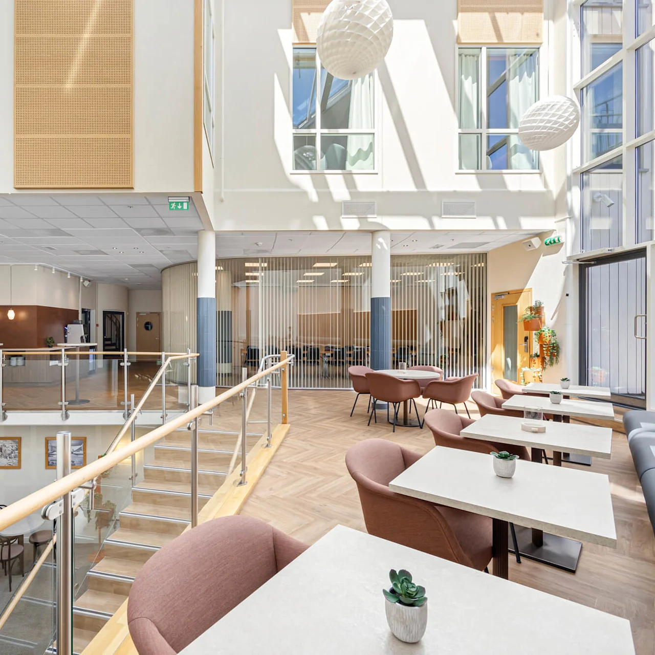 Interior of a bright, modern lobby with tables and chairs, featuring a mezzanine and tall windows allowing ample sunlight.