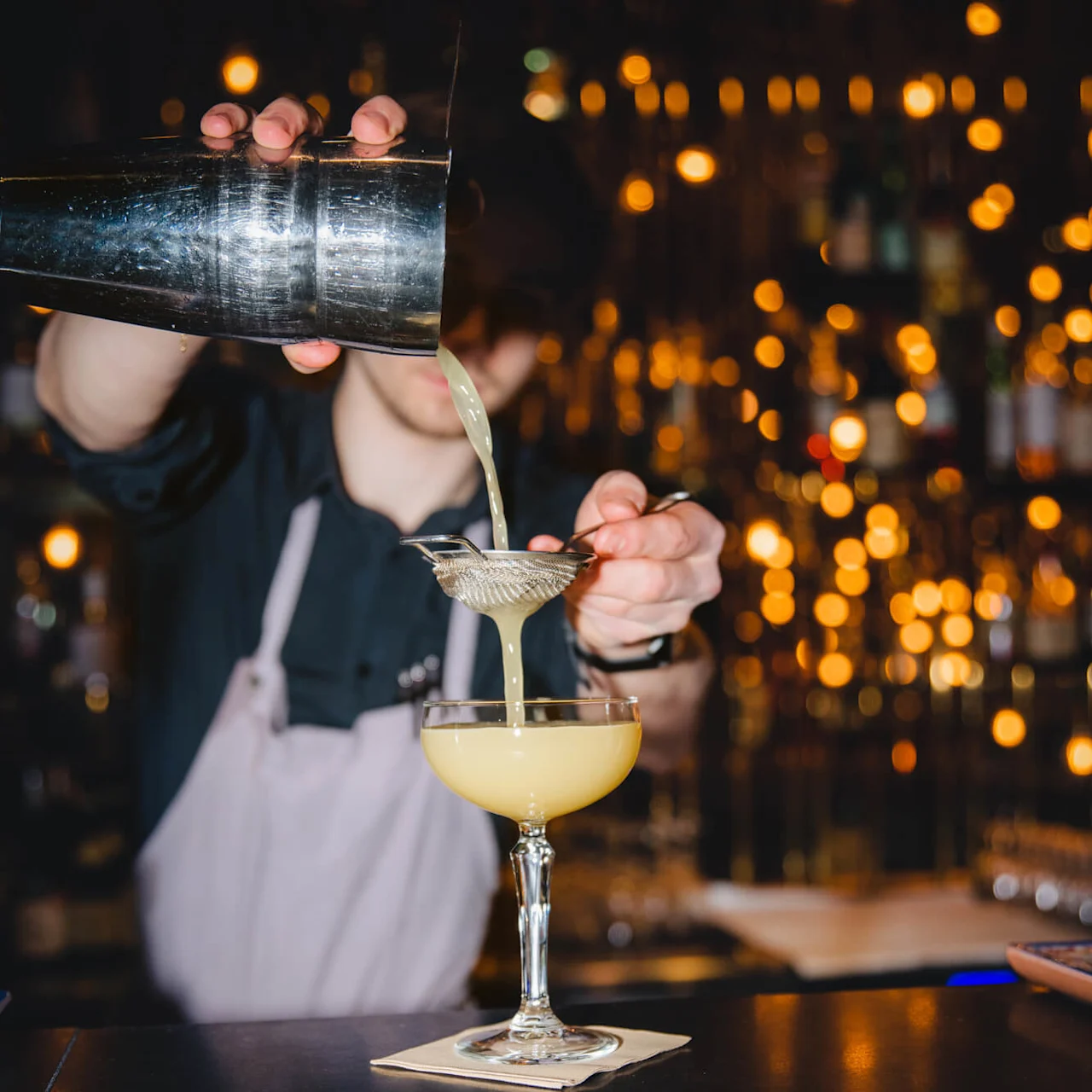 En bartender häller en gul cocktail genom en sil i ett kupat glas på en bardisk, med varma, suddiga ljus i bakgrunden.