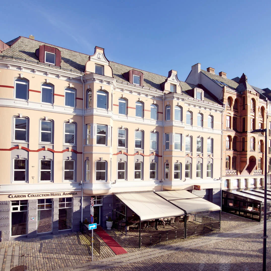 Elegante bygninger linjerer en solbelyst, brosteinsbelagt plass med markiser og noen fotgjengere, i en stille, historisk bymiljø.