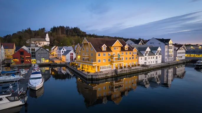 Colorful buildings line a calm waterfront, their reflections shimmering in the water under a twilight sky. Boats are docked nearby, and a forested hill rises in the background.