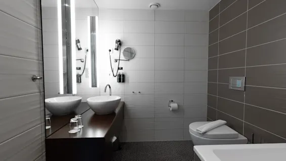 Two white sinks are set on a wooden counter beneath tall mirrors, surrounded by tiled bathroom walls, next to a toilet and bathtub in a modern, sleek design. 