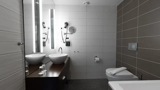 Two white sinks are set on a wooden counter beneath tall mirrors, surrounded by tiled bathroom walls, next to a toilet and bathtub in a modern, sleek design. 