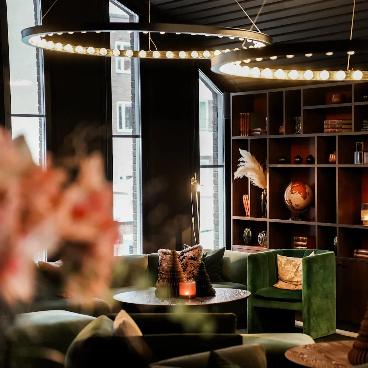 Elegant lounge med frodiga gröna stolar omger träbord, mjukt upplyst av cirkulära taklampor. Hyllor visar böcker, en jordglob och dekorativa föremål, vilket ger en mysig atmosfär.