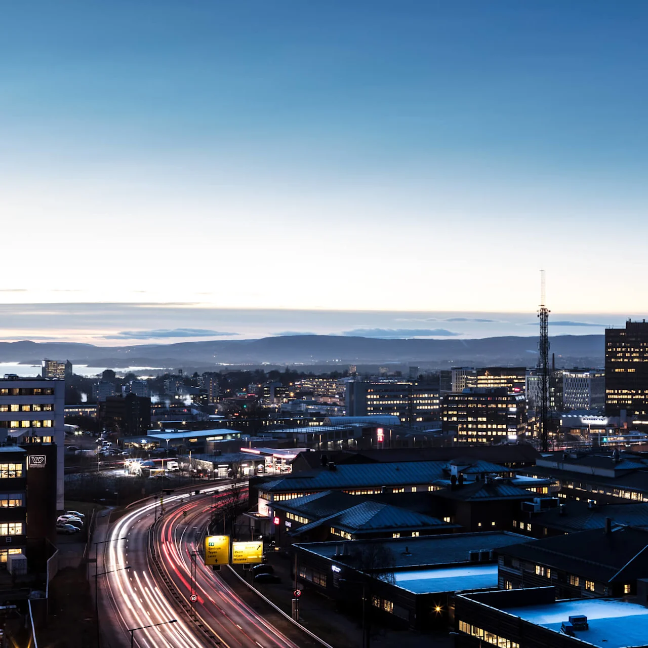 Cityscape features buildings with illuminated windows at dusk. In the foreground, light trails show moving vehicles. Mountains and water are visible in the background under a fading sky.