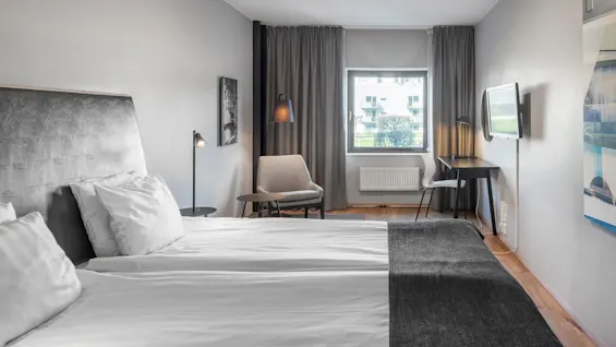 A modern hotel room features a neatly made bed, a small seating area with a chair and table, and a desk with a wall-mounted TV, all in a neutral-toned space.