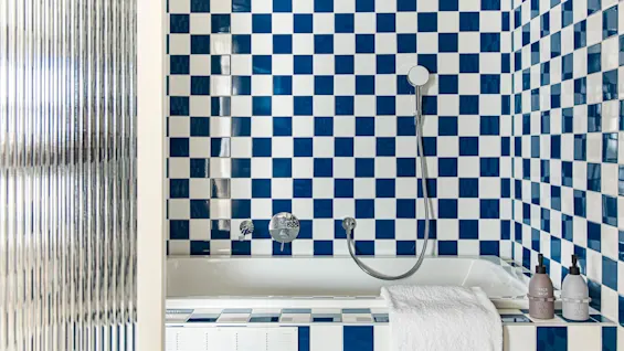 Hvidt badekar med en sølvfarvet bruser er sat mod blå og hvide ternede fliser. Der er et foldet hvidt håndklæde på kanten. To flasker med teksten "HAIR WASH" og "SOAP" står i nærheden.