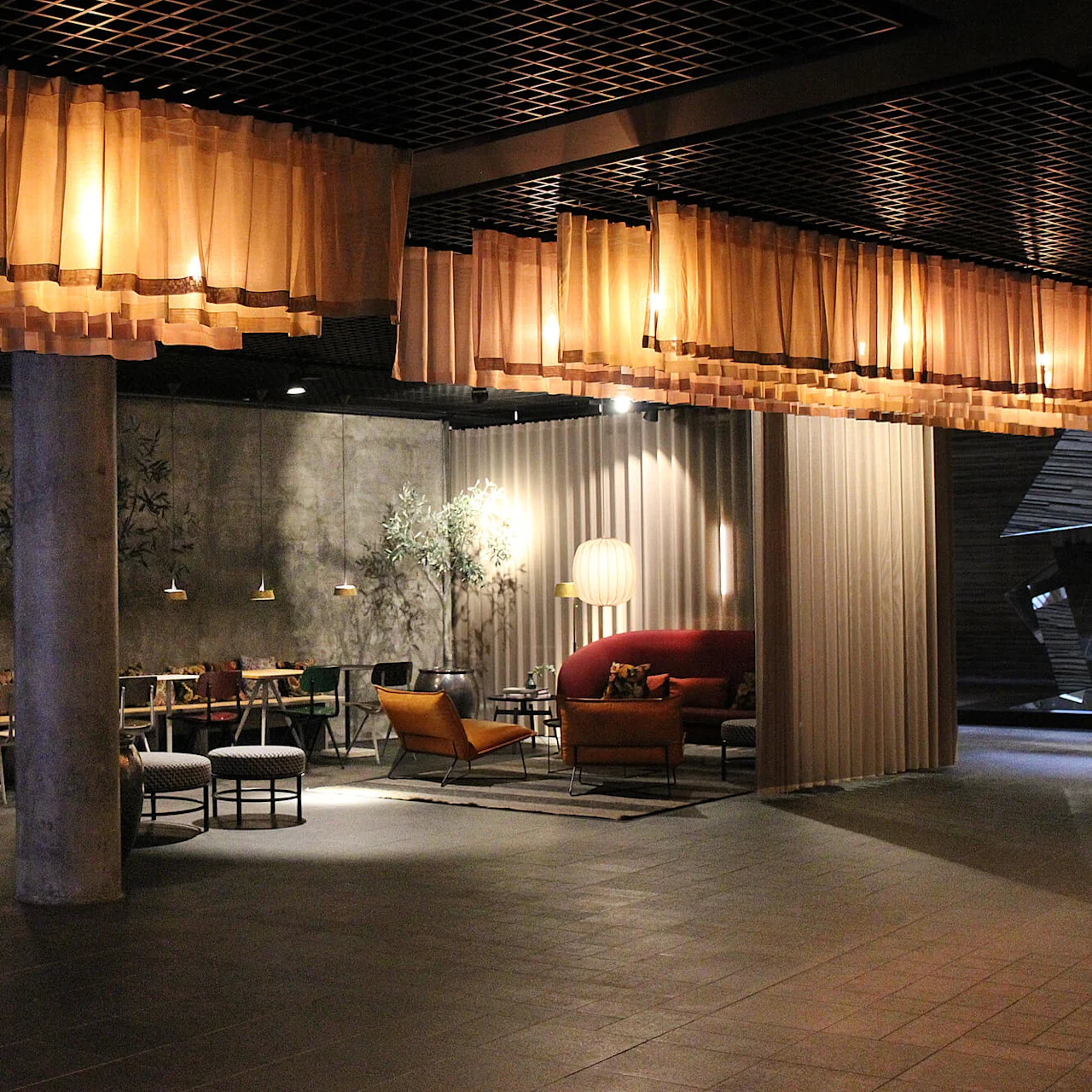 Et moderne loungeområde med hyggelige sitteplasser og varm belysning. Gardiner og planter forbedrer atmosfæren. En betongsøyle og myk lampe skaper en avslappet stemning i et stilfullt miljø.