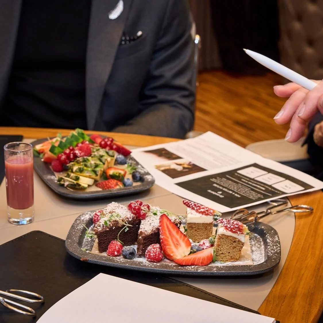 Plates of assorted pastries and fruits are on a table while people hold documents, discussing; glasses of juice and water accompany the spread in a meeting setting.