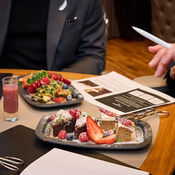 Tallrikar med olika bakverk och frukt står på ett bord medan personer håller dokument och diskuterar; glas med juice och vatten kompletterar utbudet i en mötesmiljö.