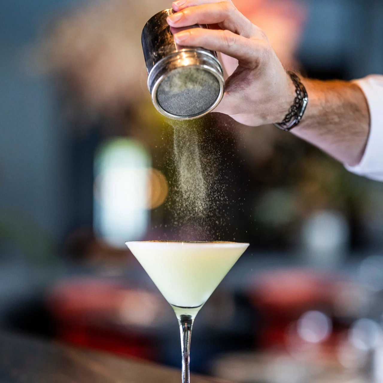 Ett cocktailglas står på ett bardisk medan en hand strör pulver från en sikta ovanför, med en suddig, färgglad bakgrund.