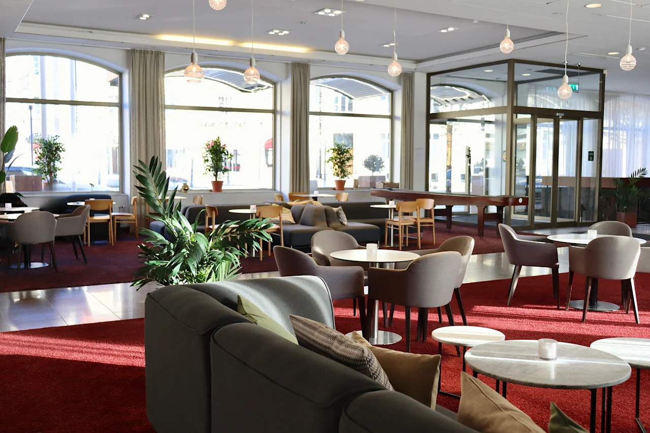 Modern hotel lobby with sofas and chairs arranged around small tables, situated on a plush red carpet. Large windows provide natural light. Potted plants add greenery, creating a welcoming atmosphere.