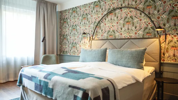 Seng med blått og hvitt teppe, mot en vegg med blomstrete mønster, opplyst av små lamper, plassert i et rom med stort vindu og myke gardiner.