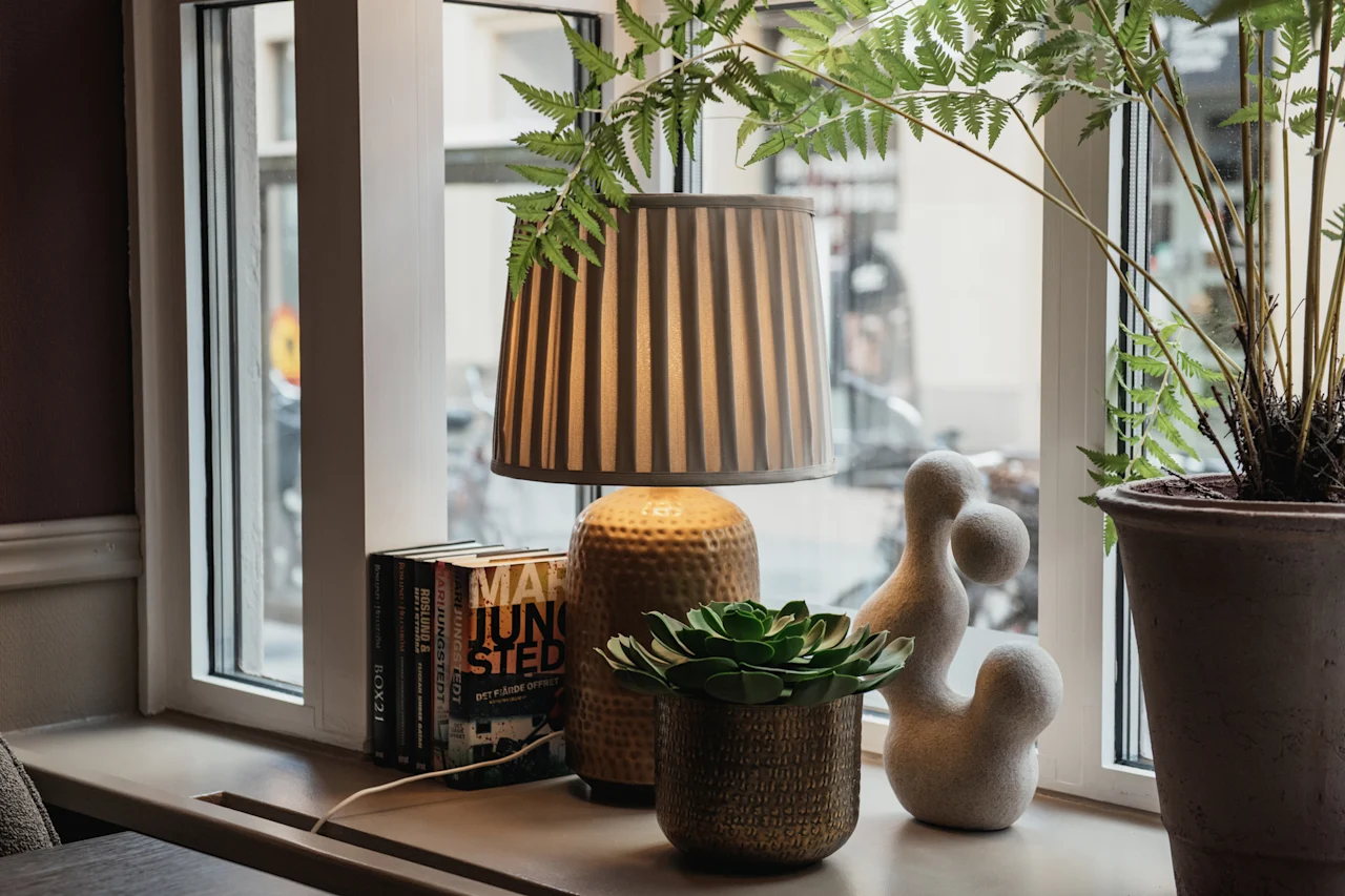 En fönsterbräda prydd med en randig lampa, böcker med titlarna MA JUNSTED och ROX21, frodiga gröna växter och en abstrakt skulptur.