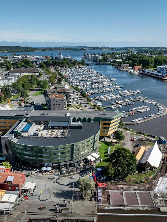 Ett böjt, modernt byggnad med gröna och gula paneler blickar ut över en småbåtshamn full med förtöjda båtar. Omkring finns bostadshus och ett färgglatt, grönt landskap mot horisonten.